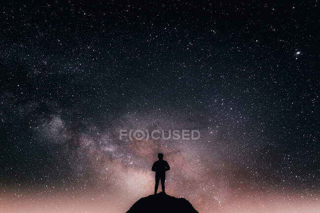 Low angle of silhouette of anonymous tourist standing on cliff against glowing starry sky at night — Stock Photo