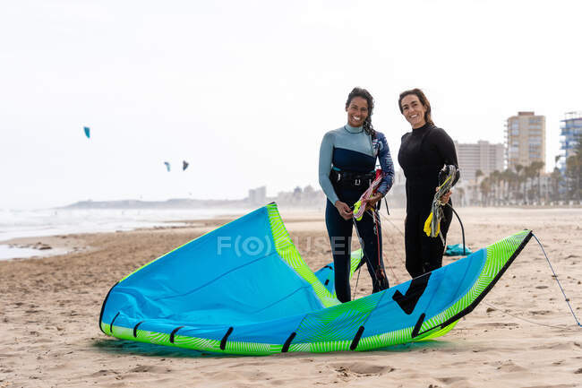 Cheerful multiethnic female kiters with power kites on sandy ocean coast under white sky in city — Stock Photo