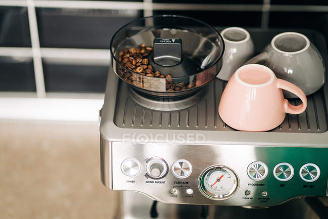 Desde arriba de granos de café tostados en molinillo eléctrico contra tazas de cerámica en rack de la moderna cafetera espresso en casa - foto de stock
