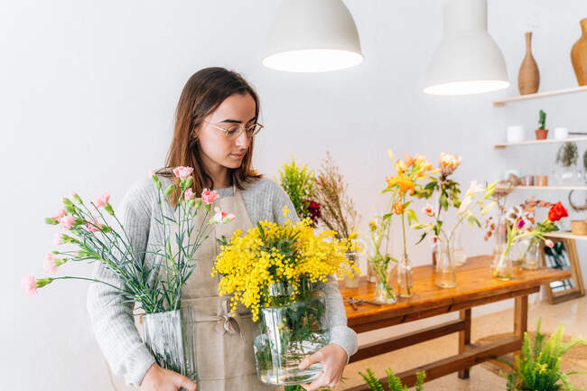 Calm young ethnic female florist in apron and eyeglasses holding glass vases with yellow mimosa and clove pink flowers in store — Stock Photo