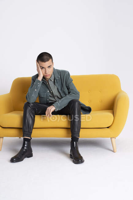 Молодой красивый мужчина-модель в модной одежде сидит на желтом диване на белом фоне в студии — стоковое фото