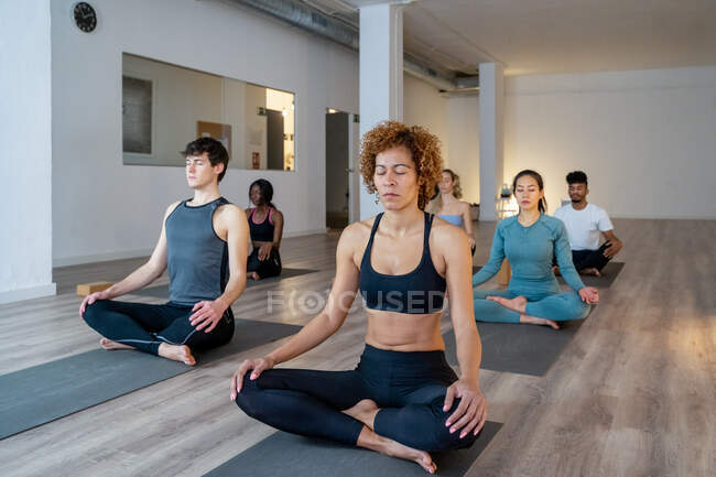 Afroamerikanerin mit einer Gruppe unterschiedlicher Menschen, die mit geschlossenen Augen in Lotus-Pose sitzt und vermittelt, während sie während des Kurses im Studio gemeinsam Yoga praktiziert — Stockfoto