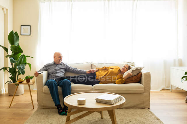 Delighted middle aged couple relaxing on comfortable couch in cozy living room while holding hands and looking at each other — Stock Photo