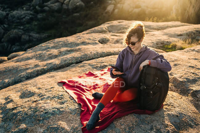 Joyful young female hiker with curly hair in casual outfit and sunglasses leaning on backpack and using smartphone while relaxing on rocky hill slope on sunny day — Stock Photo
