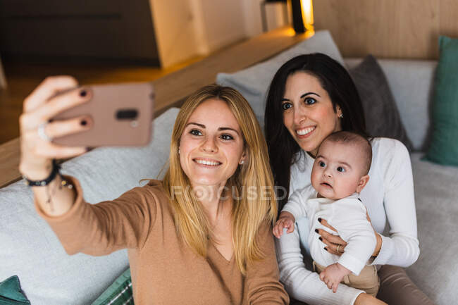 High angle of delighted LGBT family with adorable toddler sitting on couch and taking self shot on smartphone at home — Stock Photo