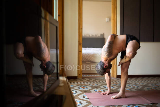 Vista lateral de un hombre sin camisa parado en Ardha Baddha Padmottanasana sobre una esterilla mientras se equilibra y practica yoga en casa - foto de stock
