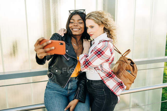Content multiethnic female friends standing on city street and taking self shot on mobile phone at weekend — Stock Photo