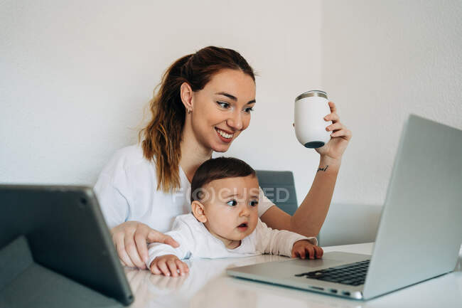 Feliz joven madre y curioso bebé en camisas blancas viendo divertido video en netbook mientras están sentados juntos en el escritorio en la sala de luz - foto de stock