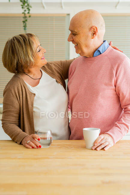 Content middle aged couple standing with cups of drinks in kitchen while hugging and looking at each other — Stock Photo