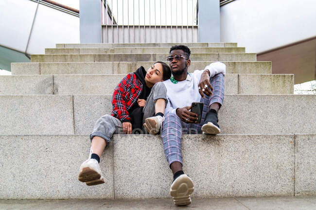 Young multiracial couple in trendy clothes sitting on city stairs with smartphone while looking away — Stock Photo