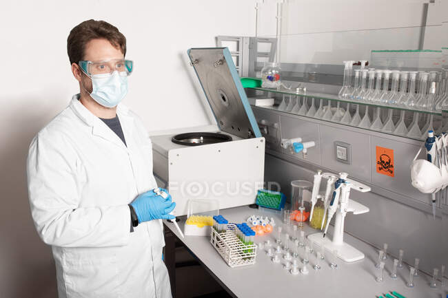 Químico masculino em máscara uniforme e descartável olhando para a frente na mesa com equipamentos profissionais e tubos de ensaio em laboratório — Fotografia de Stock