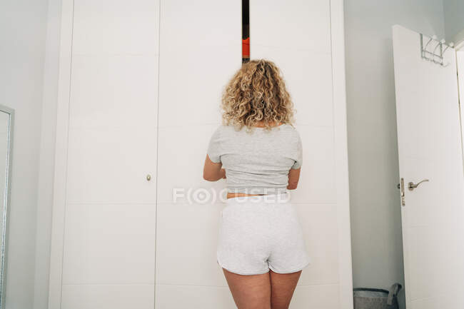 Back view of unrecognizable young woman with blond curly hair opening door of wardrobe in light apartment — Fotografia de Stock