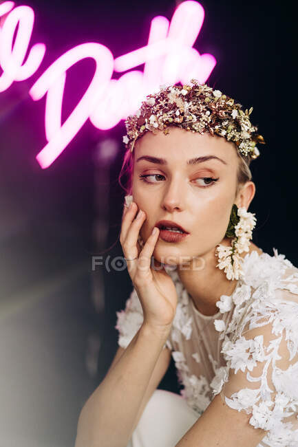 Encantadora joven novia tierna en vestido de encaje blanco y lujosa corona floral y pendientes mirando hacia otro lado contra el fondo negro con luces de neón - foto de stock