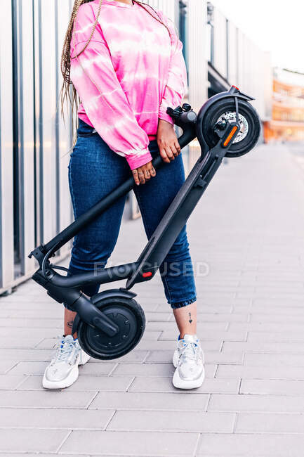 Crop anonymous female in casual wear standing with modern scooter on tiled walkway in town — Stock Photo