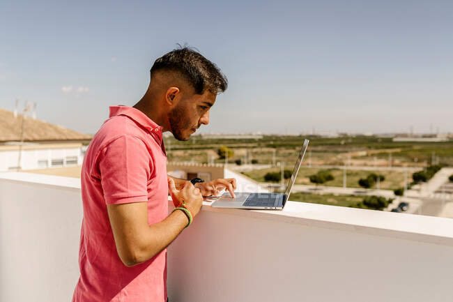 Сторона зору сфокусованого молодого етнічного чоловічого фрілансера з темним волоссям в тренді, стоячи на терасі і використовуючи ноутбук під час дистанційної роботи. — стокове фото