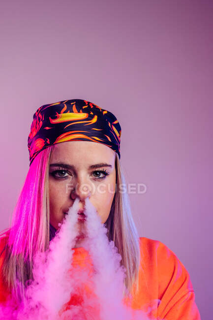 Fêmea legal em roupa de estilo de rua fumando e cigarro e exalando fumaça pelo nariz em fundo roxo no estúdio com iluminação de néon rosa — Fotografia de Stock