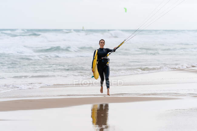 Female athlete in wetsuit with control bar looking at camera on sandy shore against foamy ocean after practicing kiteboarding — Stock Photo