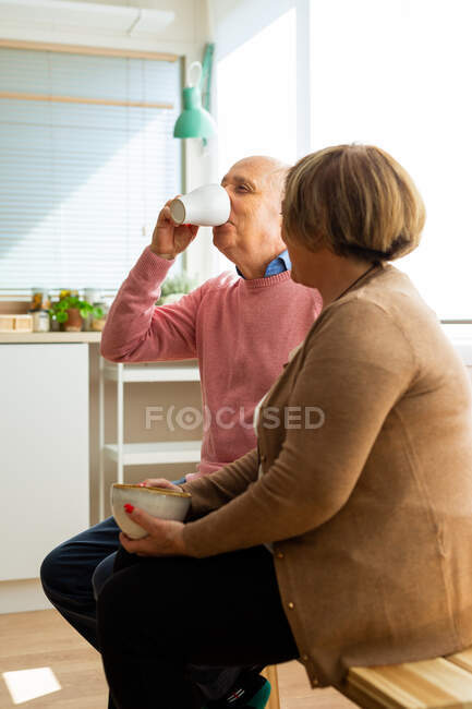 Middle aged couple sitting on wooden table in spacious kitchen and drink coffee — Stock Photo