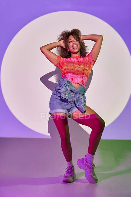Ganzkörperfitte afroamerikanische Tänzerin in kurzen Hosen, die mit erhobenen Armen tanzt und die Zunge herausstreckt, um wegzuschauen, während sie im Studio im Neonlicht steht — Stockfoto