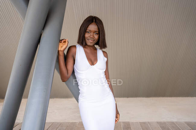 Feliz atraente afro-americano fêmea vestindo elegante vestido branco com decote baixo em pé perto de postes de concreto na rua e olhando para a câmera — Fotografia de Stock