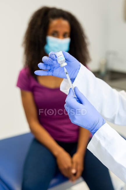 Обрезанный неузнаваемый врач в латексных перчатках, наполняющий шприц из бутылки вакциной для вакцинации молодой афроамериканской пациентки в клинике во время вспышки коронавируса — стоковое фото