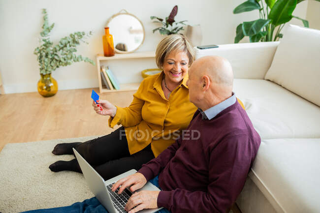 Munteres älteres Paar, das beim Online-Einkauf zu Hause per Laptop mit Plastikkarte bezahlt — Stockfoto
