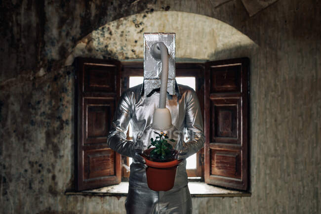 Unerkennbare Person in silbernem Kostüm mit Atemschutzgerät und Schlauch an Topfpflanze befestigt steht in schäbigem verlassenen Raum — Stockfoto