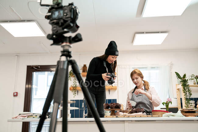 Женщина фотографирует шоколадные кексы на цифровой камере против блогера, разговаривающего во время приготовления пищи на кухне — стоковое фото