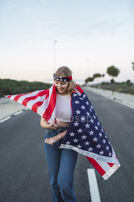 Encantada mujer americana de pie envuelta con la bandera nacional de EE.UU. en la carretera al atardecer y mirando hacia abajo - foto de stock