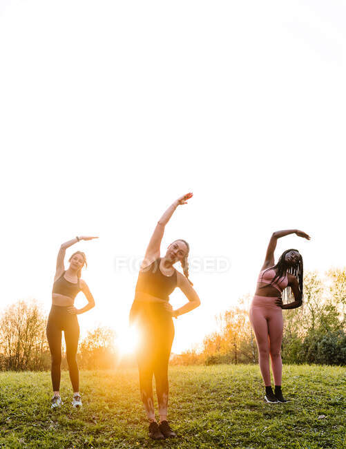 Female athletes doing side bend exercise while stretching together in park on background of sunset sky — Stock Photo