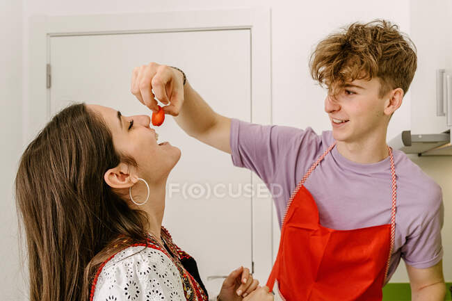 Joven alegre en ropa casual y delantal alimentando a la feliz novia étnica con fresa mientras cocinan juntos en la cocina - foto de stock