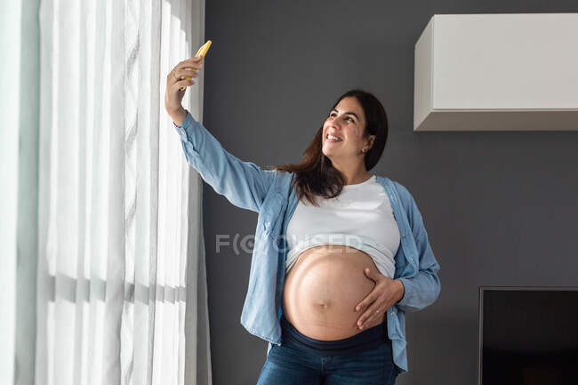 Sorrindo fêmea grávida tocando barriga enquanto estava no quarto em casa e tomando selfie com telefone celular — Fotografia de Stock