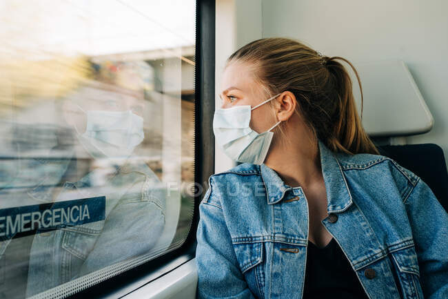Mujer joven en chaqueta de mezclilla y máscara facial protectora mirando hacia otro lado en la ventana del tren mientras viaja - foto de stock