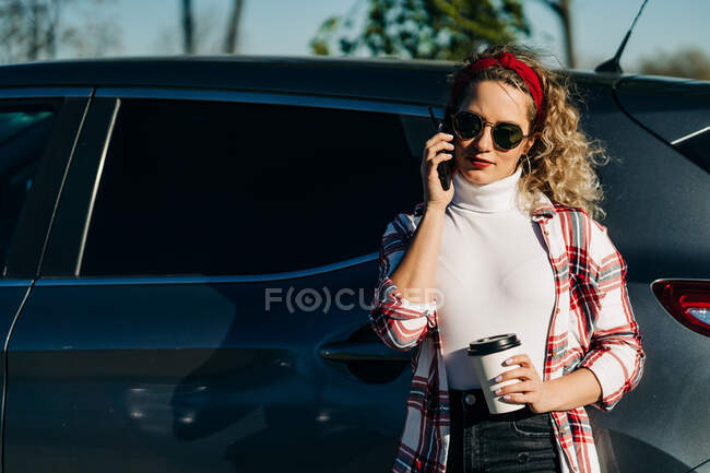 Mujer de moda de pie con bebida para llevar cerca de un automóvil moderno y hablando por teléfono móvil - foto de stock