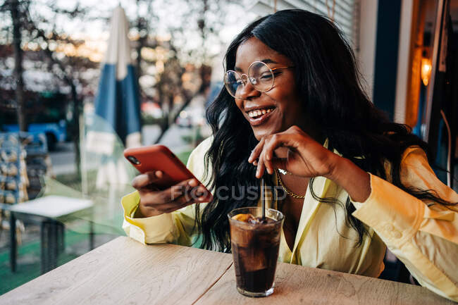 Femme afro-américaine élégante assise à table dans un café avec boisson rafraîchissante soda et la navigation sur les médias sociaux sur le téléphone mobile — Photo de stock