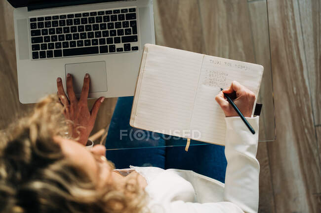 Зверху анонімна студентка з кучерявим волоссям у повсякденному одязі, що сидить за скляним столом і робить нотатки в копіювальній книзі, готуючись до іспитів, використовуючи ноутбук вдома — стокове фото