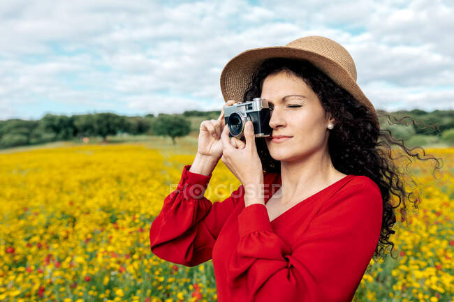 Усміхнена жінка в капелюсі фотографується на старовинній камері на лузі під хмарним небом — стокове фото