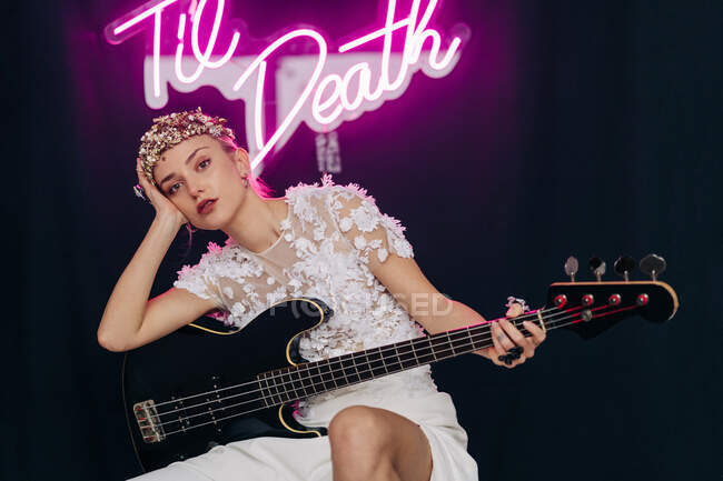 Energetic rebellious young woman in elegant white bridal dress and wreath with guitar in hand making horn gesture in studio with neon inscription — Foto stock