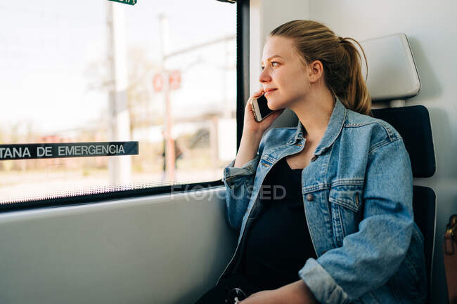 Молода випадкова жінка в джинсовій куртці має телефонний дзвінок, дивлячись у вікно поїзда під час роботи — стокове фото