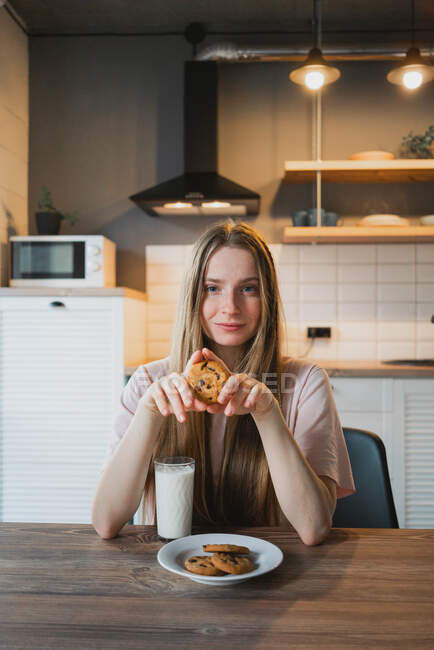 Alegre joven hembra con sabrosa galleta de avena con chips de chocolate para el desayuno en la mesa en la cocina - foto de stock