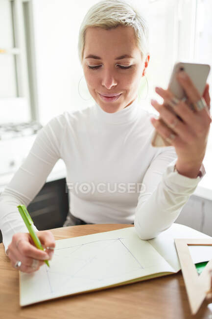 Astrologue souriante avec des lignes de dessin de téléphone portable dans l'album papier à la table dans la maison légère — Photo de stock