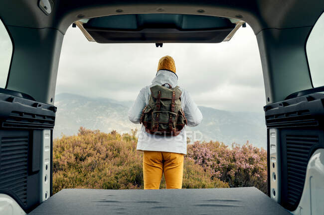 Back view of male camper in outerwear standing near van and admiring scenic view of highlands — Stock Photo