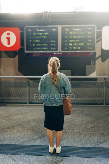 Rückenansicht einer Frau in Jeansjacke und mit Tasche beim Blick auf den Fahrplan der Züge an Bord, die auf dem Bahnhof stehen — Stockfoto