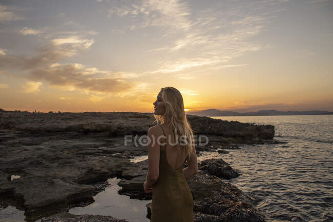 Vue latérale de la jeune femme debout sur la côte contre la mer bleu ondulant au coucher du soleil et regardant loin — Photo de stock