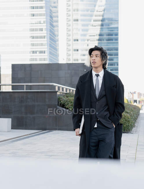 Молодой этнический мужчина исполнительный в пальто с рукой в кармане и нетбук глядя в сторону во время прогулки в городе — стоковое фото