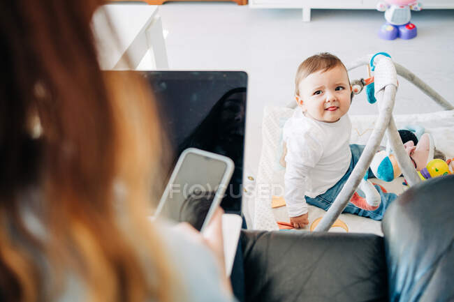 Liebenswertes kleines Baby, das mit Spielzeug auf dem Boden sitzt und die zugeschnittene anonyme Mutter beim Surfen auf dem Handy im hellen Wohnzimmer ansieht — Stockfoto