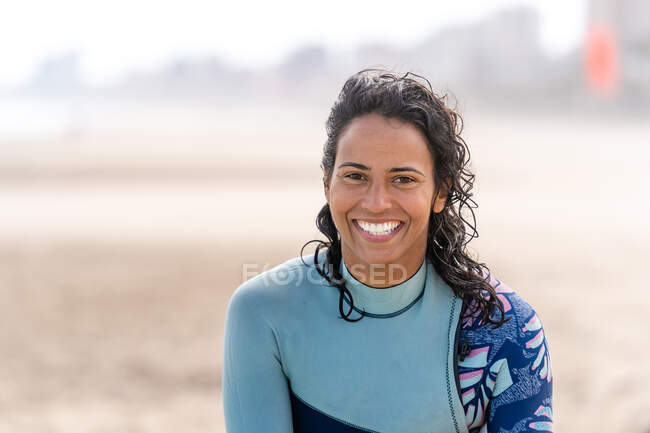 Happy ethnique femelle kiter en combinaison avec équipement de kitesurf regardant la caméra sur la plage de sable de l'océan — Photo de stock