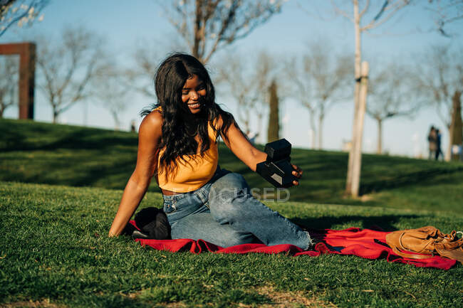 Encantada hembra afroamericana tomando fotos en cámara fotográfica instantánea retro mientras está sentada en una manta en el parque en la noche de verano - foto de stock