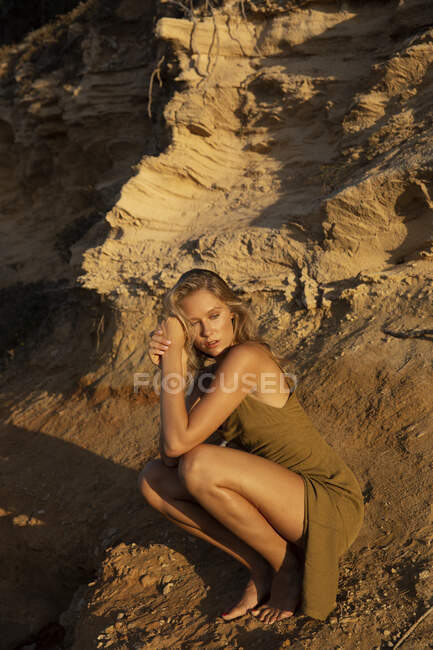 Corpo inteiro de mulher loira sensual descalça sentada em assombrações perto de encosta arenosa e olhando para longe — Fotografia de Stock