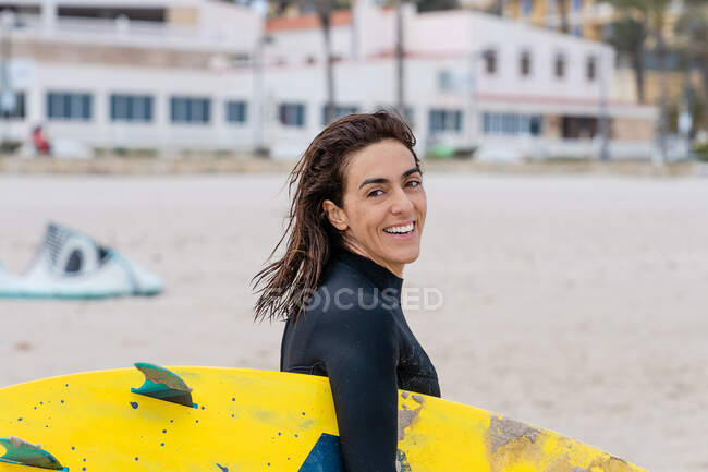 Side view of cheerful adult female kitesurfer with wet hair and kiteboard looking at camera on sandy beach — Stock Photo